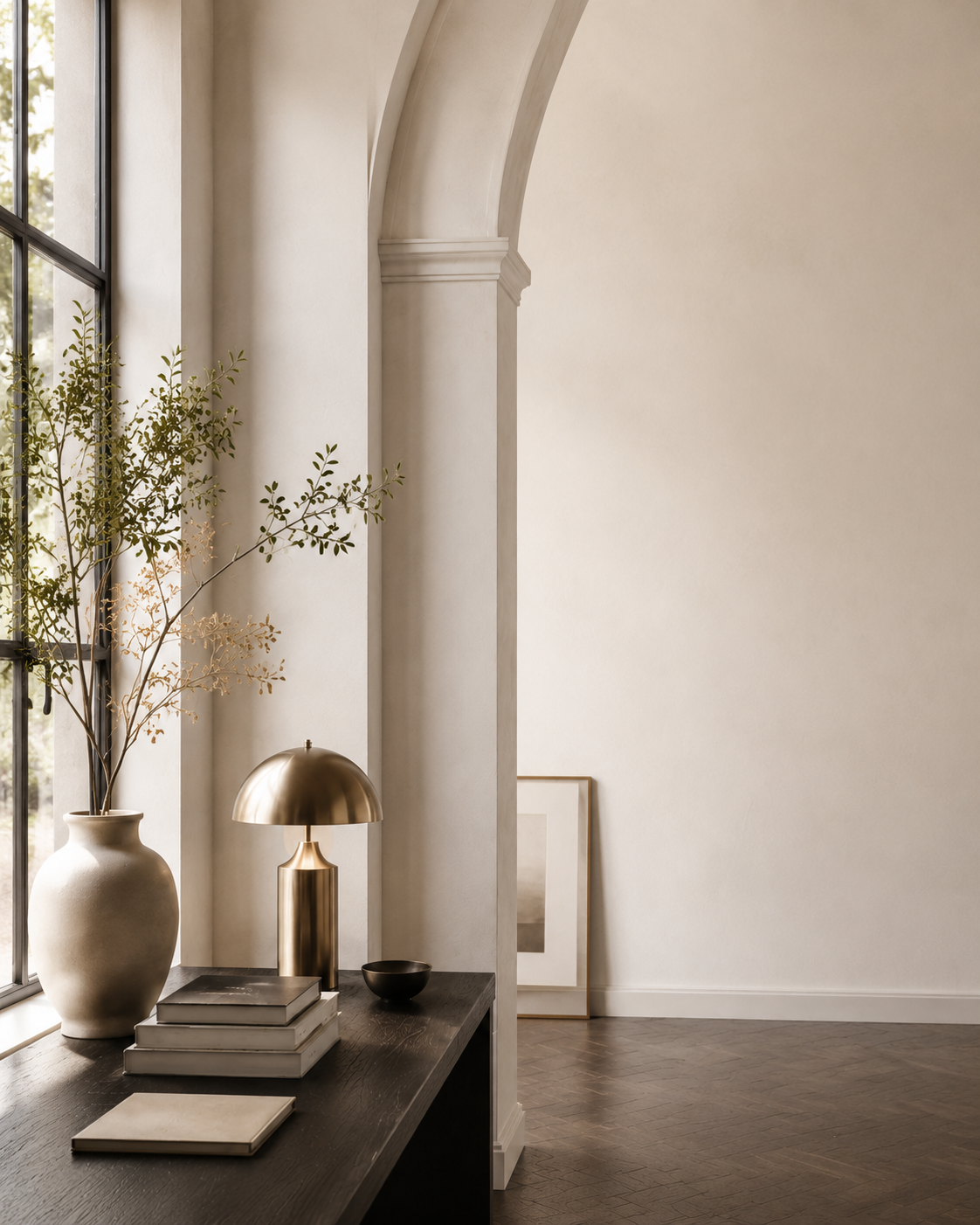 Warm, sunlit studio interior with an arched wall, a leafy branch, and a brass desk lamp.