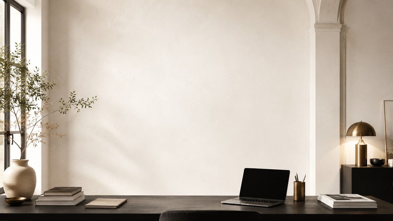 Warm editorial workspace with textured plaster, a black desk, books, a laptop, and brass accents